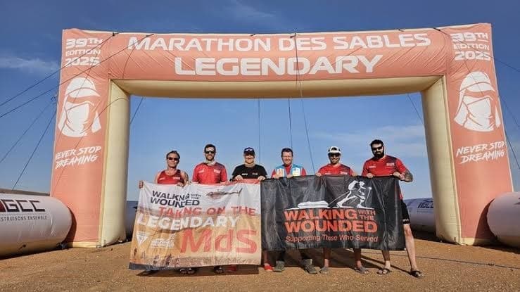 Six Walking With The Wounded runners holding team banners under the Marathon des Sables Legendary arch, 2025